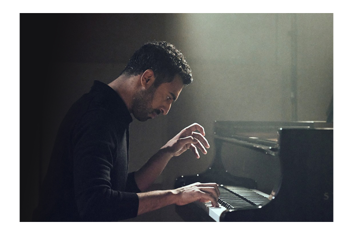 A man wearing a black jumper playing the piano under a spotlight.